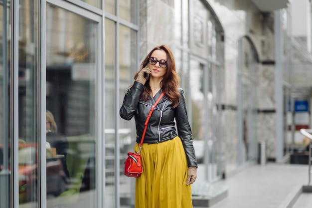 Frau mit der roten Handtasche, die beim Sprechen am Telefon nahe einem Geschäftsgebäude geht