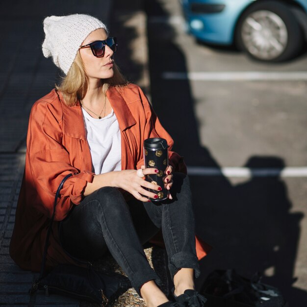 Frau mit der Thermosflasche, die auf Pflasterung sitzt