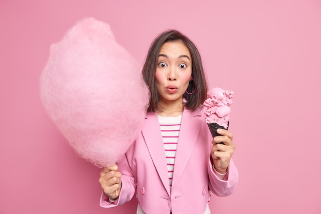 Frau mit dunklem Haar hat sich gefragt, Gesichtsausdruck hält Kegeleis und Zuckerwatte, gekleidet in formelle elegante Kleidung