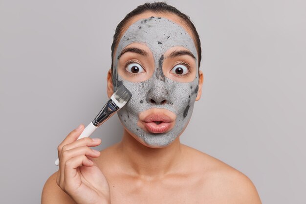 Frau mit dunklem Haar trägt Tonpeeling-Maske auf Gesicht hält Kosmetikpinsel starrt mit großem Staunen auf Kamerabesuche Spa-Salon steht oben ohne auf grauer Wand