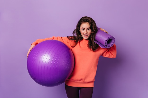 Frau mit dunklem voluminösem Haar im orangefarbenen Sweatshirt hält fitball und Fitnessmatte auf lila Wand