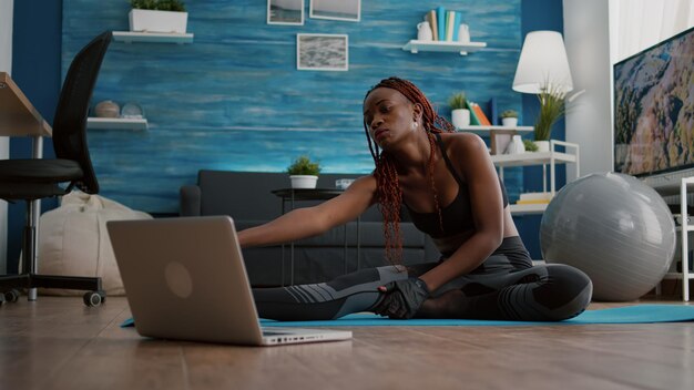 Frau mit dunkler Haut, die Pilates-Training im Wohnzimmer macht und Körpermuskeln auf Yoga-Karte streckt, während sie Online-Fitness-Sportvideos auf dem Laptop sieht. Flexibler Erwachsener, der einen gesunden Lebensstil genießt
