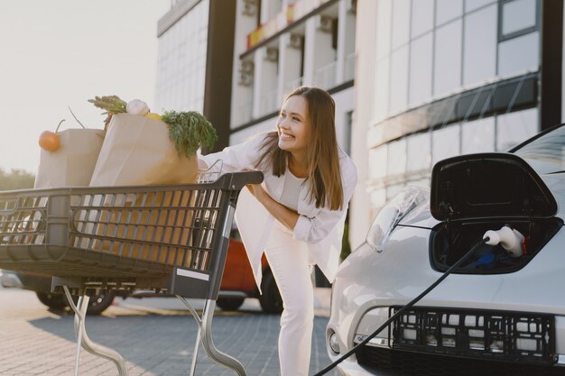 Frau mit einem Einkaufswagen, der Elektroauto an der elektrischen Tankstelle auflädt