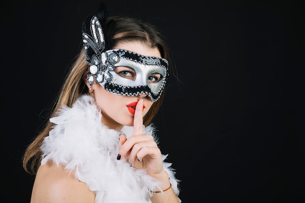Frau mit einer Karnevalsmaske, die Ruhegeste auf schwarzem Hintergrund macht