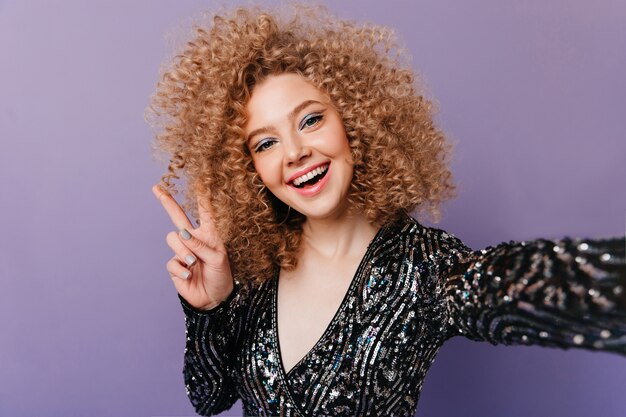 Frau mit glänzenden Schatten und blonden Locken, Top in Pailletten tragend, zeigt Friedenszeichen und macht Selfie auf lila Raum.