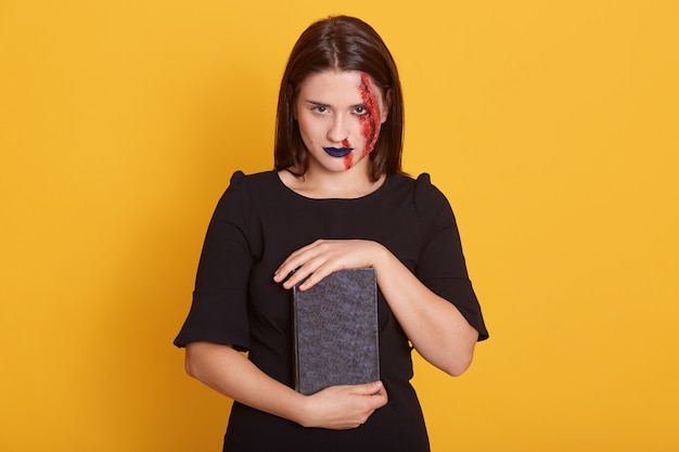 Frau mit Horror Halloween Make-up und blutige Wunde posiert im Studio auf gelb, junge Frau mit demgerous Anblick hält Buch mit Beschwörung, Kleider schwarzes Kleid