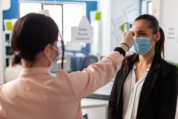 Frau mit Infrarot-Thermometer zur Messung der Temperatur von Büroangestellten während der globalen Epidemie mit Coronavirus in einem Geschäftsunternehmen. Neue Normalität in Zeiten der Weltpandemie mit Covid19.