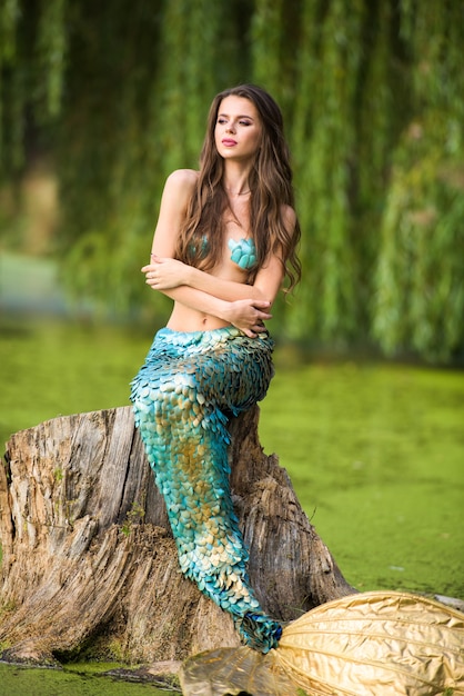 Frau mit langen braunen Haaren und gekleidet wie eine Meerjungfrau sitzt auf dem Stein über Wasser