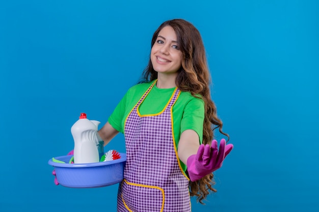 Frau mit langen gewellten Haaren, die Schürze und Gummihandschuhe tragen, die Becken voll von Reinigungswerkzeugen halten, die willkommene Geste lächelnd freundlich stehend auf Blau machen