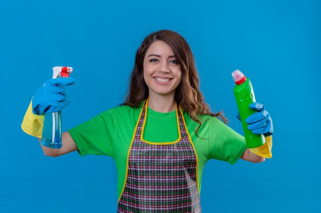 Frau mit langen gewellten Haaren, die Schürze und Gummihandschuhe tragen und Flaschen mit Reinigungsmitteln in erhobenen Händen halten, die fröhlich auf blau stehend lächeln