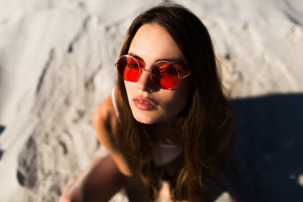Frau mit langen Haaren in roten Sonnenbrillen sitzt auf weißem Sand