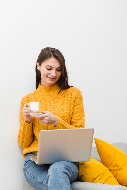 Frau mit Laptop auf dem Schoss, der Tasse Kaffee beim Sitzen auf dem Sofa hält