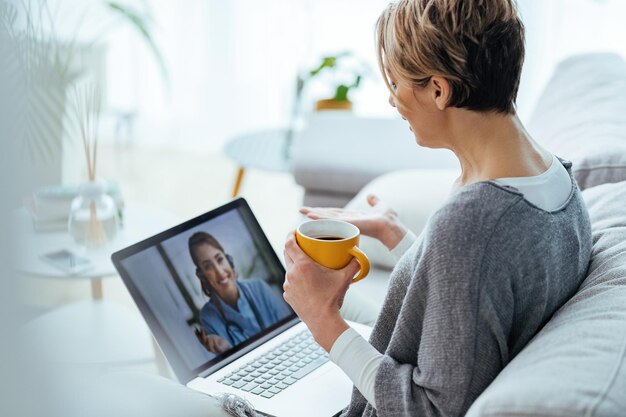 Frau mit Laptop und Videoanruf mit ihrem Arzt, während sie zu Hause sitzt