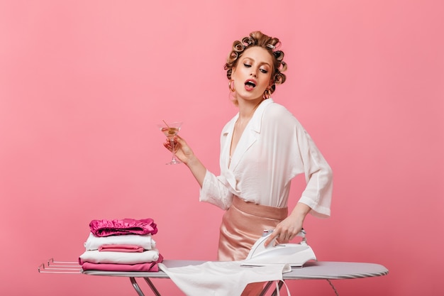 Frau mit Locken gekleidet in eleganten Kleidern, die Martini-Glas halten und Leinen bügeln