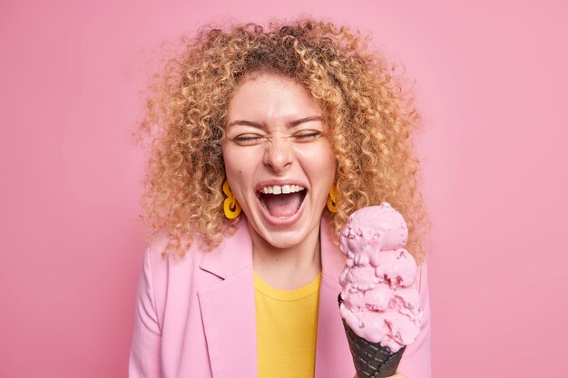 Frau mit lockigem, buschigem Haar hat Spaß, leckeres Eis isst gerne leckeres gefrorenes Dessert in modischer Jacke