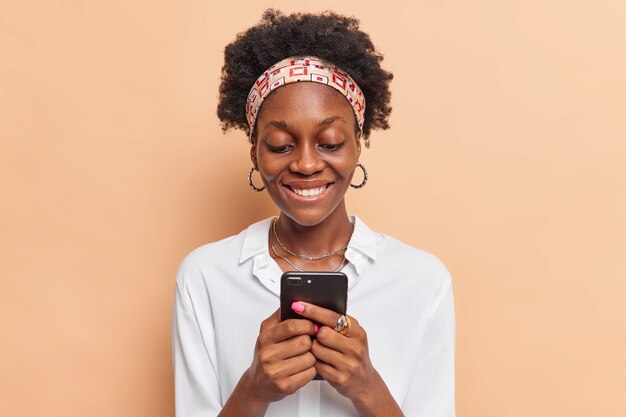 Frau mit lockigem Haar benutzt Handy-Chats online bekommt Nachricht von Freund trägt Stirnbandohrringe weißes Hemd isoliert auf Beige
