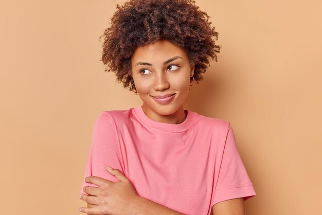 Frau mit lockigem Haar schaut weg und ist tief in Gedanken versunken trägt lässiges rosa T-Shirt isoliert auf Beige erinnert an schöne Erinnerung. Menschen und Gedanken Konzept