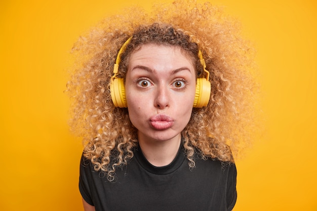Frau mit lockigem Haar sieht mit kokettem Ausdruck in die Kamera hält die Lippen gefaltet hört Musik über Kopfhörer genießt die Freizeit in lässigen T-Shirt-Posen