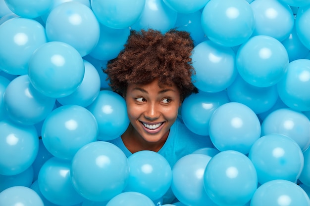 Frau mit lockigem Haar streckt den Kopf durch aufgeblasene blaue Luftballons aus lächelt breit hat fröhliche Stimmung feiert etwas.