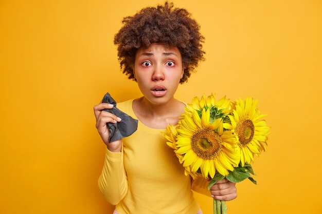 Frau mit lockigen Haaren, roten, geschwollenen Augen, hält Serviettenniesen wegen Sonnenblumenallergie, die krank ist, posiert drinnen auf leuchtendem Gelb