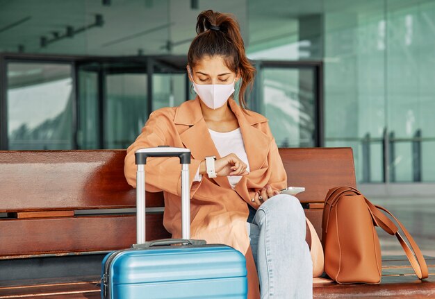 Frau mit medizinischer Maske, die ihre Uhr am Flughafen während der Pandemie betrachtet