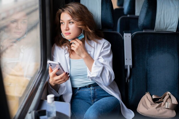 Frau mit medizinischer Maske, die mit dem öffentlichen Zug reist und Smartphone verwendet