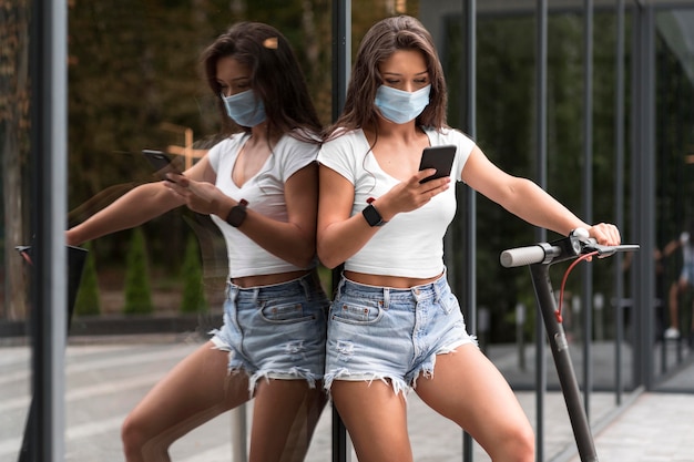 Frau mit medizinischer Maske, die Smartphone neben Elektroroller prüft
