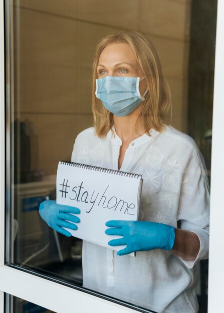 Frau mit medizinischer Maske halten zu Hause Notizbuch am Fenster