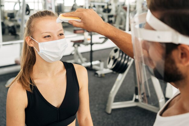 Frau mit medizinischer Maske im Fitnessstudio, die ihre Temperatur vom Mann mit Gesichtsschutz überprüfen lässt