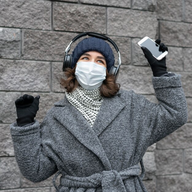 Frau mit medizinischer Maske in der Stadt, die Musik mit Kopfhörern und Smartphone hört