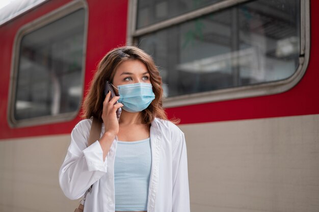 Frau mit medizinischer Maske mit Smartphone am öffentlichen Bahnhof