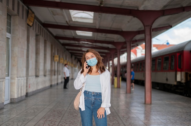 Frau mit medizinischer Maske mit Smartphone am öffentlichen Bahnhof