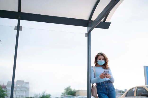 Frau mit medizinischer Maske mit Smartphone am öffentlichen Busbahnhof