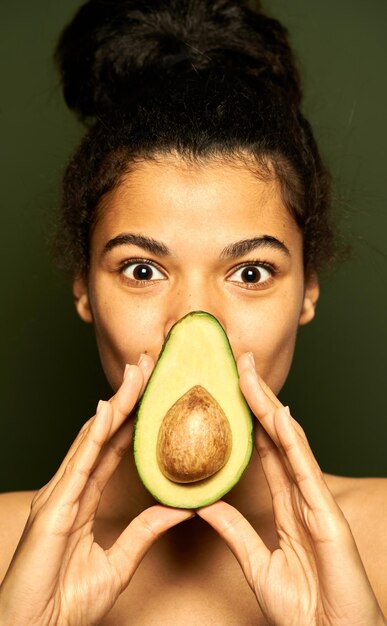Frau mit perfekter Haut, die in die Kamera schaut, Avocado halb auf dem Mund hält und isoliert auf Grün posiert