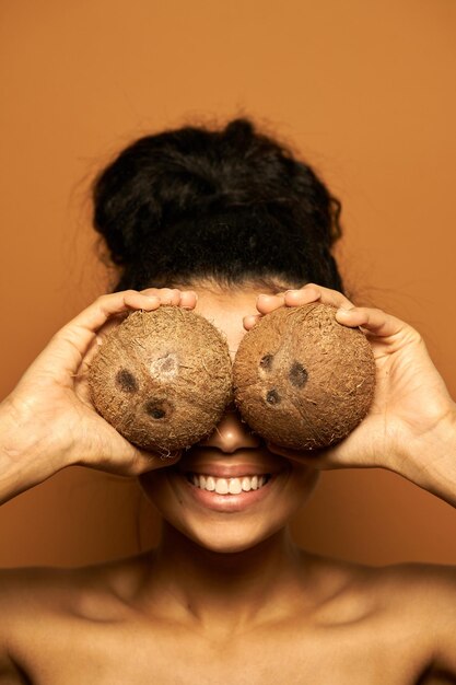 Frau mit perfekter Haut und Haaren in einem Brötchen versteckt ihre Augen hinter zwei Kokosnüssen und posiert isoliert auf Orange