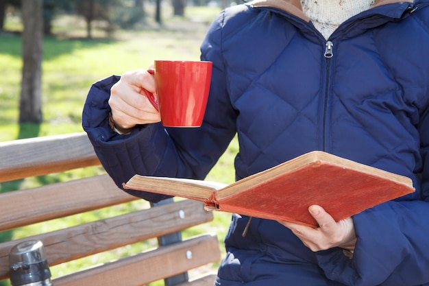 Frau mit roter tasse kaffee und altem buch, sitzend auf einer bank im stadtpark.