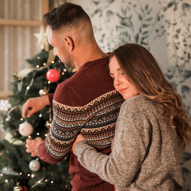 Frau mit schließenden Augen, die Mann von der Rückseite in den Strickjacken umarmen, nähern sich Weihnachtsbaum