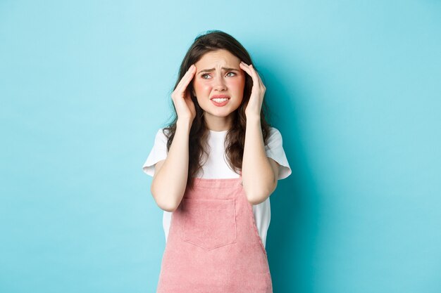 Frau mit schmerzhafter Migräne, Kopf berühren und Stirnrunzeln, Blick in die obere linke Ecke, Kopfschmerzen, stehend auf blauem Hintergrund