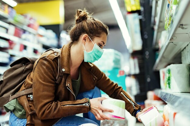 Frau mit schützender Gesichtsmaske, die während der Virusepidemie Toilettenpapier im Supermarkt kauft