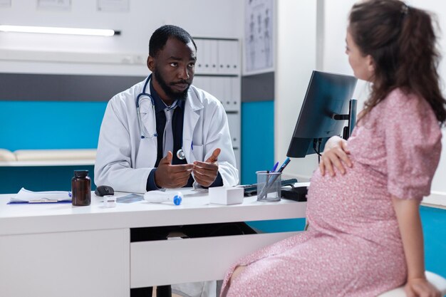 Frau mit Schwangerschaft, die mit einem Spezialisten über die medizinische Versorgung im Büro diskutiert. Allgemeinmediziner, der mit Patienten spricht, die ein Kind erwarten, über Gesundheitsversorgung und Unterstützung, das Tragen von Gesichtsmasken