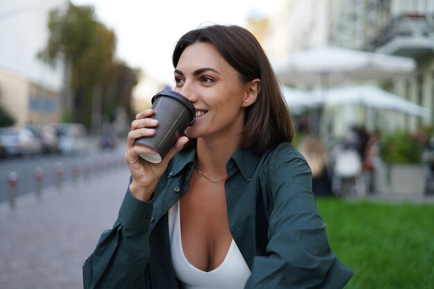 Frau mit Tasse Kaffee im Freien an der Stadtstraße bei Sonnenuntergang glücklich lächelnd genießen Sommertage