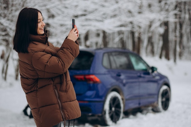 Kostenloses Foto frau mit telefon steht an ihrem auto im winterwald