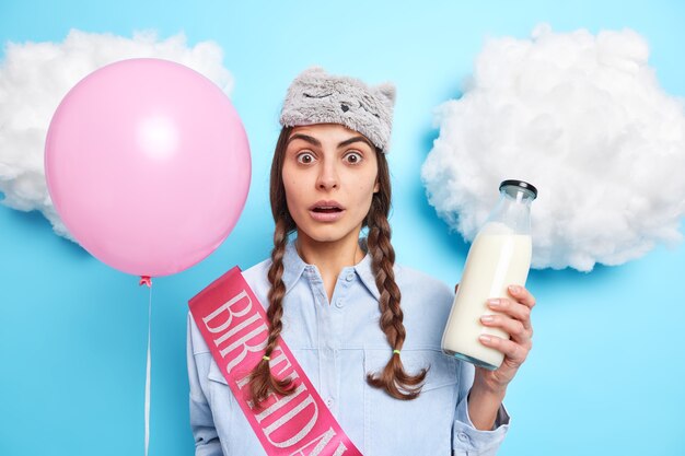 Frau mit zwei Zöpfen hält rosa aufgeblasenen Ballon und Glasflasche Milch trägt Band mit Aufschrift Geburtstag isoliert auf Blau