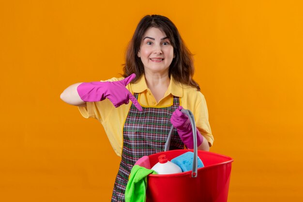 Frau mittleren Alters, die Schürze und Gummihandschuhe trägt, die Eimer mit Reinigungswerkzeugen halten, die mit dem Finger darauf zeigen, positiv und glücklich lächelnd über orange Wand 3 stehend