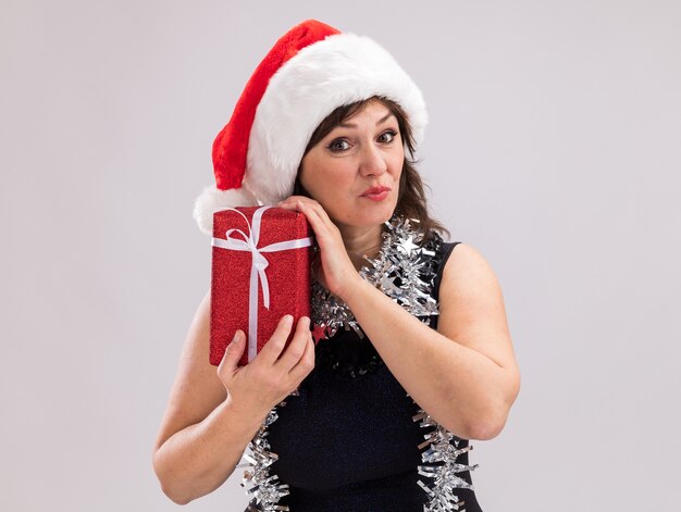 Frau mittleren Alters, die Weihnachtsmütze und Lametta-Girlande um den Hals hält, hält Weihnachtsgeschenkpaket, das Kamera lokalisiert auf weißem Hintergrund mit Kopienraum betrachtet