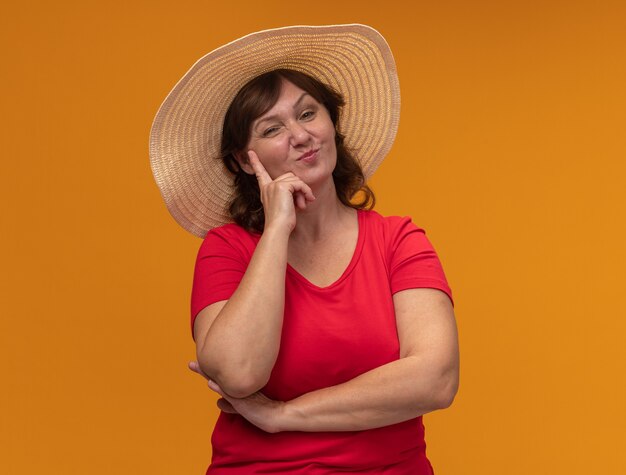 Frau mittleren Alters im roten T-Shirt und im Sommerhut mit dem glücklichen Gesicht lächelnd, das über orange Wand steht