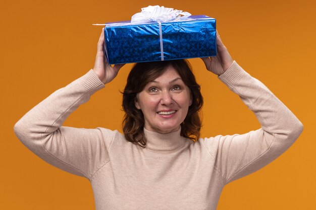 Frau mittleren Alters in beigem Rollkragenpullover, der Geschenkbox über ihrem Kopf glücklich und aufgeregt hält über orange Wand hält