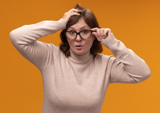 Frau mittleren Alters in beigem Rollkragenpullover mit Brille überrascht über orange Wand stehend