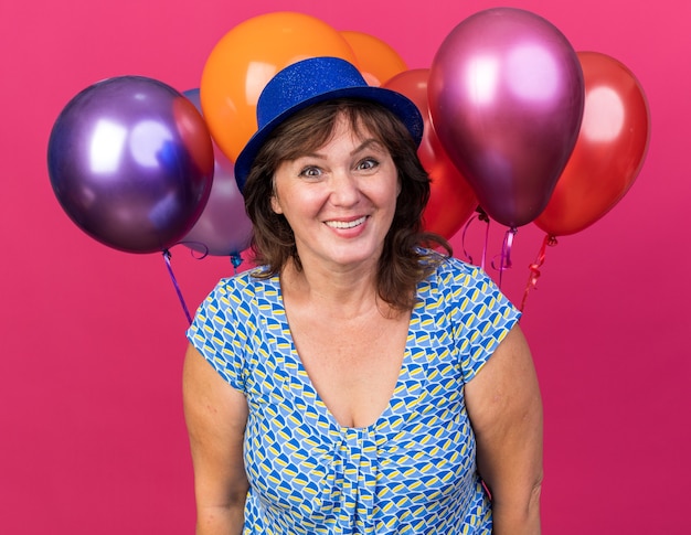 Frau mittleren Alters in Partyhut, die bunte Luftballons mit glücklichem Gesicht hält, die fröhlich lächelt und die Geburtstagsfeier feiert, die über rosa Wand steht