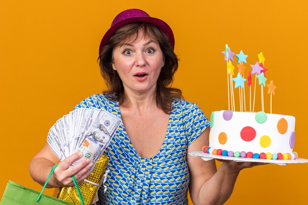 Frau mittleren Alters in Partyhut, die Papiertüte mit Geburtstagsgeschenken und Kuchen hält, erstaunt und überrascht, die Geburtstagsfeier über orangefarbener Wand zu feiern?